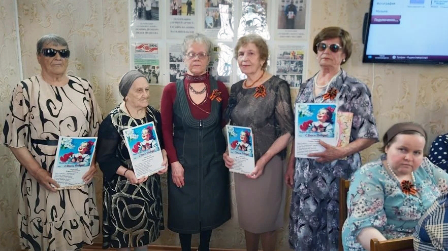 На фото шесть женщин пожилого возраста в помещении. Пять стоят с грамотами в руках и георгиевскими ленточками на груди, одна сидит справа в цветочном платье.