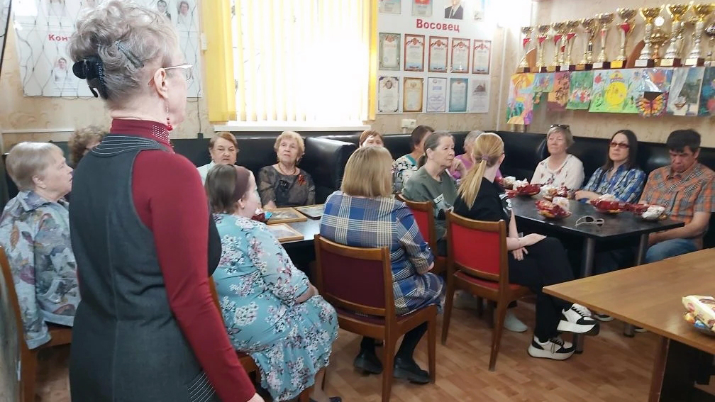 Группа людей, в основном пожилые женщины, сидят за столами в комнате с кубками и детскими рисунками на стене.
Перед ними стоит женщина в бордовой водолазке и тёмном сарафане