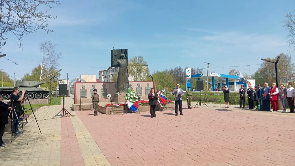 высокий памятник с венками, слева памятный танк, у монумента выступают люди с микрофоном, справа стоит группа зрителей.