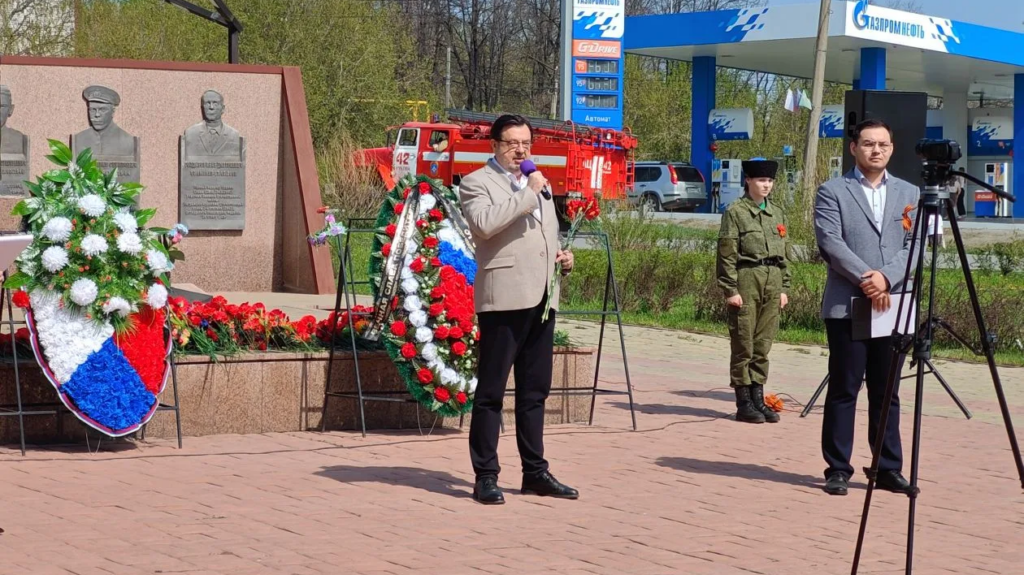 Торжественное мероприятие у военного памятника: мужчина выступает с микрофоном, рядом ведущий и девушка в форме, на фоне венки, пожарная машина и заправка «Газпромнефть».