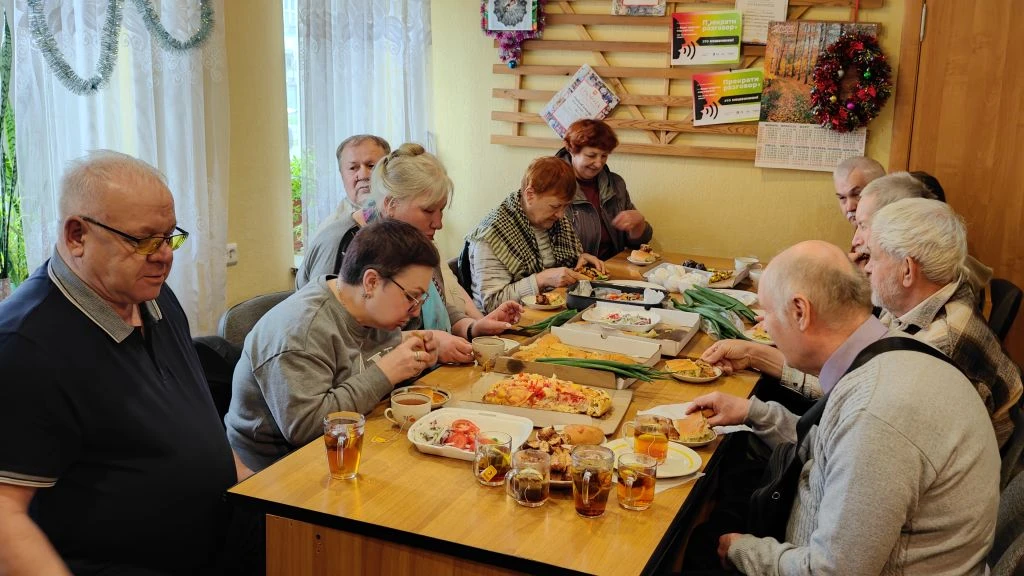 За большим столом сидит группа пожилых людей, светлокожих, и вместе ест: на столе пицца, закуски, хлеб, чай в стаканах и кружках.