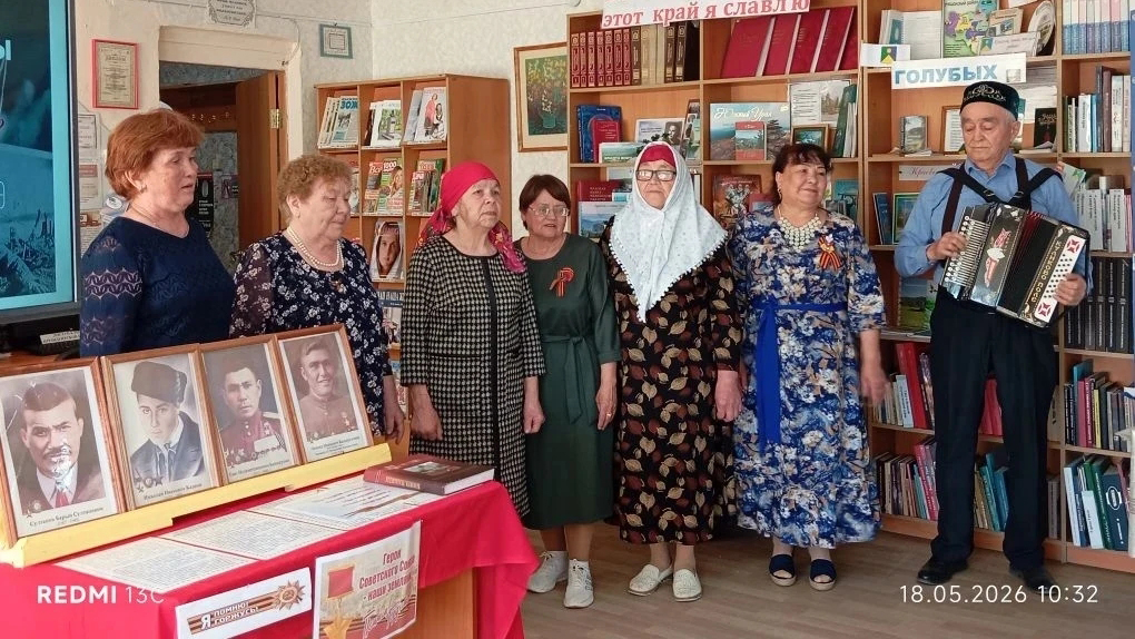 Группа пожилых женщин и мужчина с гармошкой поют или выступают в библиотеке. Слева стол с портретами фронтовиков, сзади книжные стеллажи.