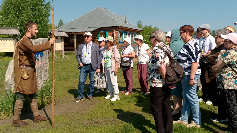 Группа пожилых людей на экскурсии на улице слушает молодого мужчину в старинной одежде с длинным посохом, на фоне деревянного дома.
