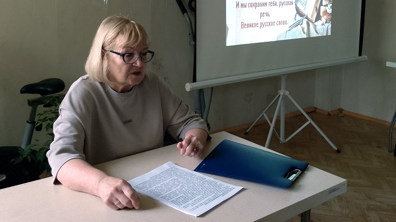 Светловолосая женщина в очках сидит за столом, перед ней лист с текстом и синяя папка; на фоне — экран проектора с слайдом и текстом про русскую речь.