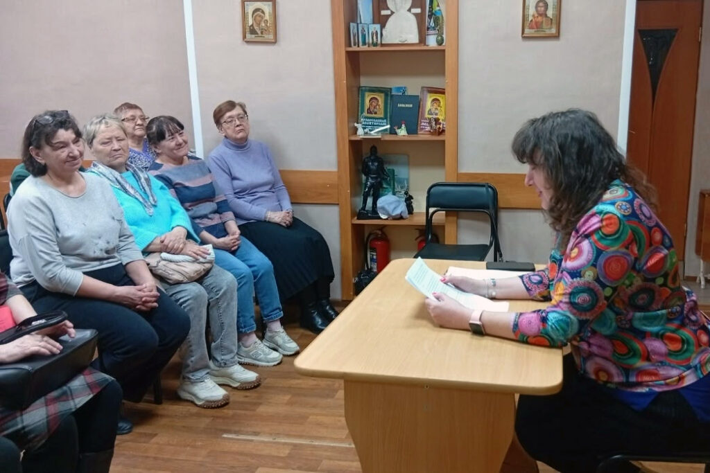 Женщина в яркой разноцветной блузке сидит за столом и читает текст с листа.
Напротив неё на стульях сидит группа пожилых женщин, внимательно слушают; за ними стеллаж с иконами и книгами.
