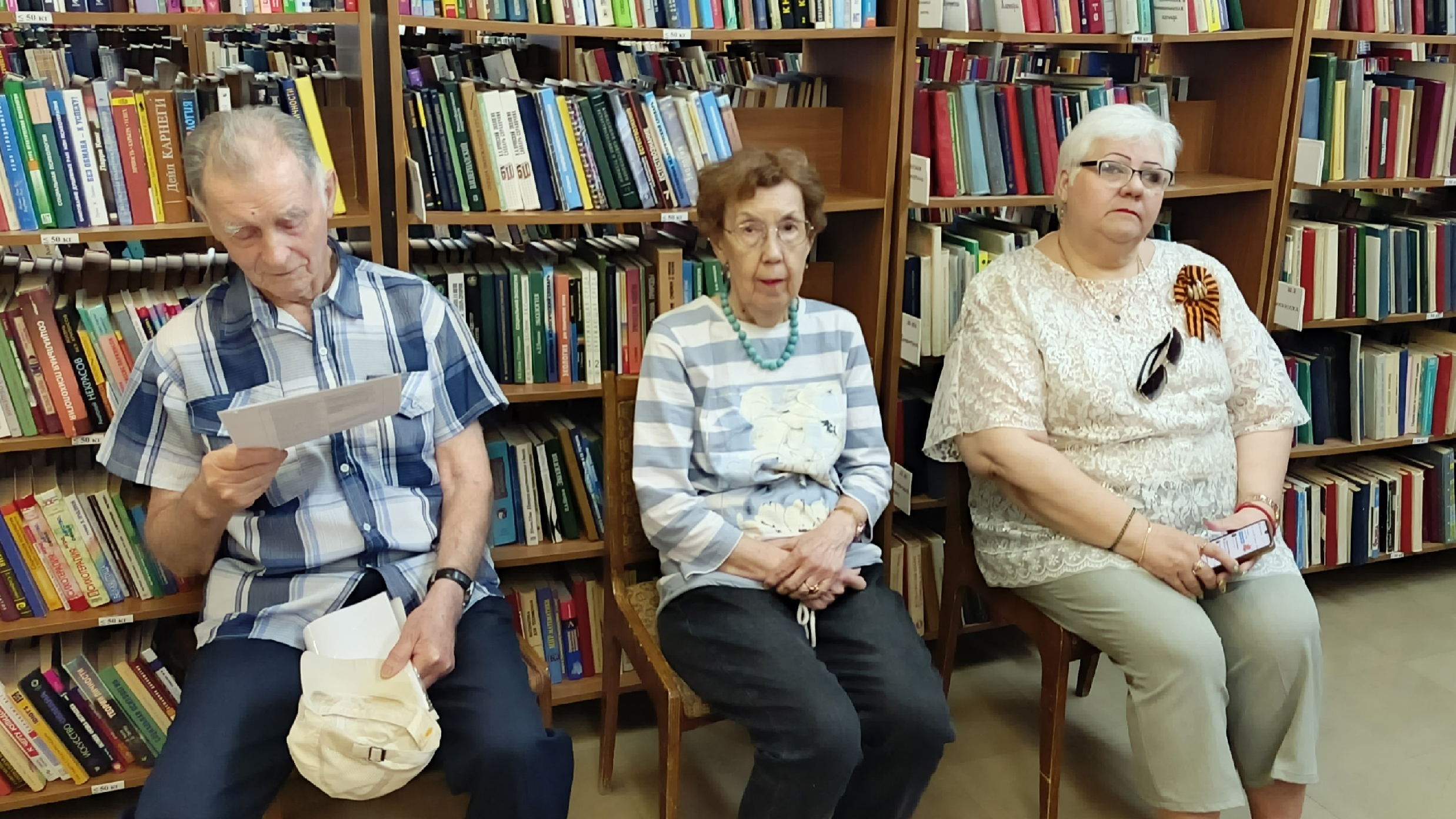 Три пожилых человека сидят на стульях в библиотеке перед книжными полками. Слева мужчина читает лист бумаги. Посередине женщина в полосатой кофте с бирюзовым ожерельем. Справа женщина в кружевной блузке с георгиевской лентой и телефоном в руках.