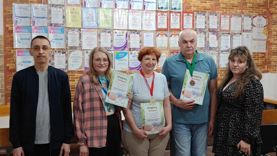 Пять человек стоят в ряду на фоне стены с грамотами; трое в центре в медалях на шее держат в руках грамоты, по краям — мужчина и женщина без наград.