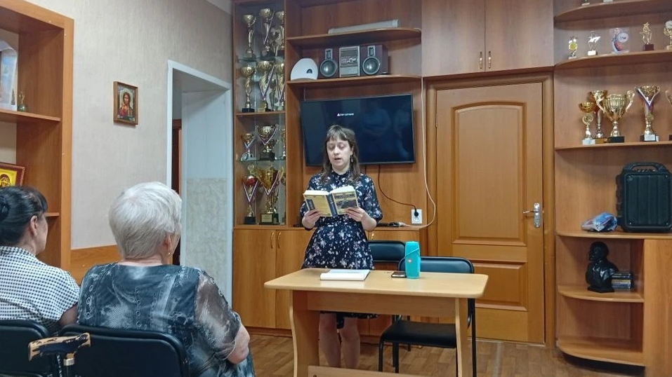 Молодая женщина стоит за столом в комнате с кубками и медалями, держит раскрытую книгу и читает вслух сидящим перед ней людям.