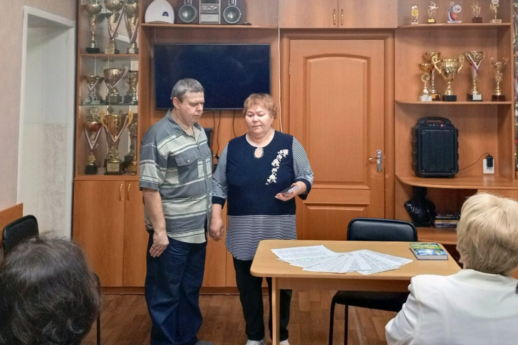 перед сидящими слушателями стоят мужчина в полосатой рубашке и женщина в темном платье с узором; на столе перед ними разложены листы с текстом и книга.