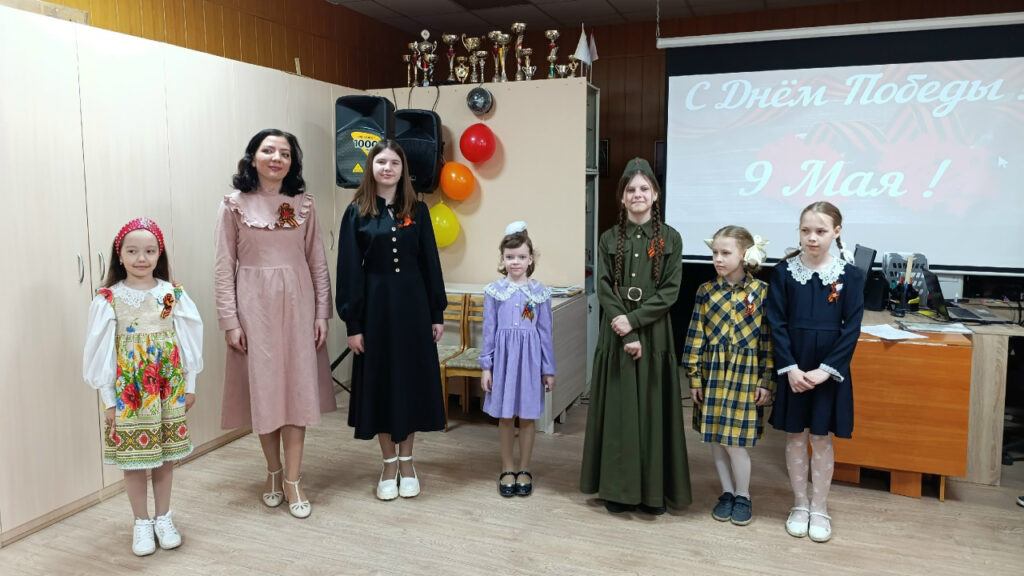 На сцене в ряд стоят семь человек: взрослая женщина и шесть девочек в нарядных и стилизованных под военное время платьях, у всех приколоты георгиевские ленточки.
Сзади экран с надписью «С Днём Победы! 9 Мая!», рядом шкаф с кубками и несколько воздушных шаров.