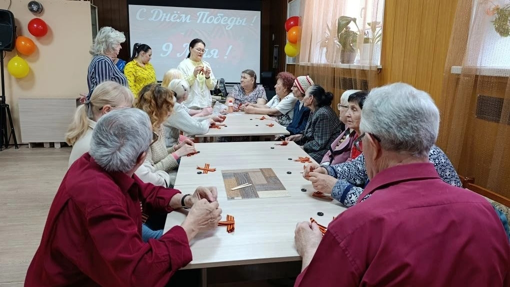Комната с длинным столом, по сторонам сидят пожилые люди и мастерят георгиевские ленточки.
В конце стола стоят ведущие, на экране сзади надпись: «С Днём Победы! 9 Мая!», вокруг разноцветные шары.