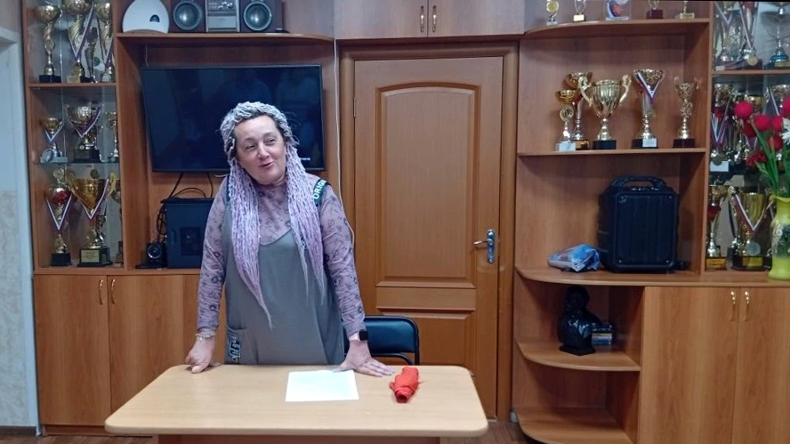 Женщина стоит за небольшим столом с листом бумаги и свёрнутой красной тканью, позади шкафы с многочисленными кубками и медалями и выключенный телевизор.