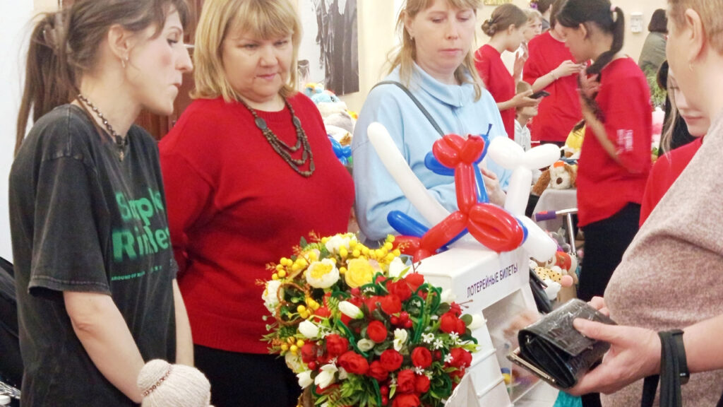 Женщины у стола с букетом и фигурками из шаров, рядом ящик «ЛОТЕРЕЙНЫЕ БИЛЕТЫ» на ярмарке.
