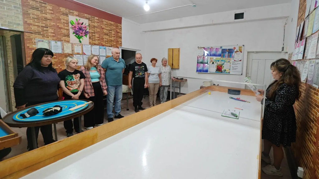Группа людей стоит у длинного стола для настольного тенниса слепых, напротив них женщина с бумагами в руках, судья, объясняет или подводит итоги в спортивном зале с грамотами на стенах.