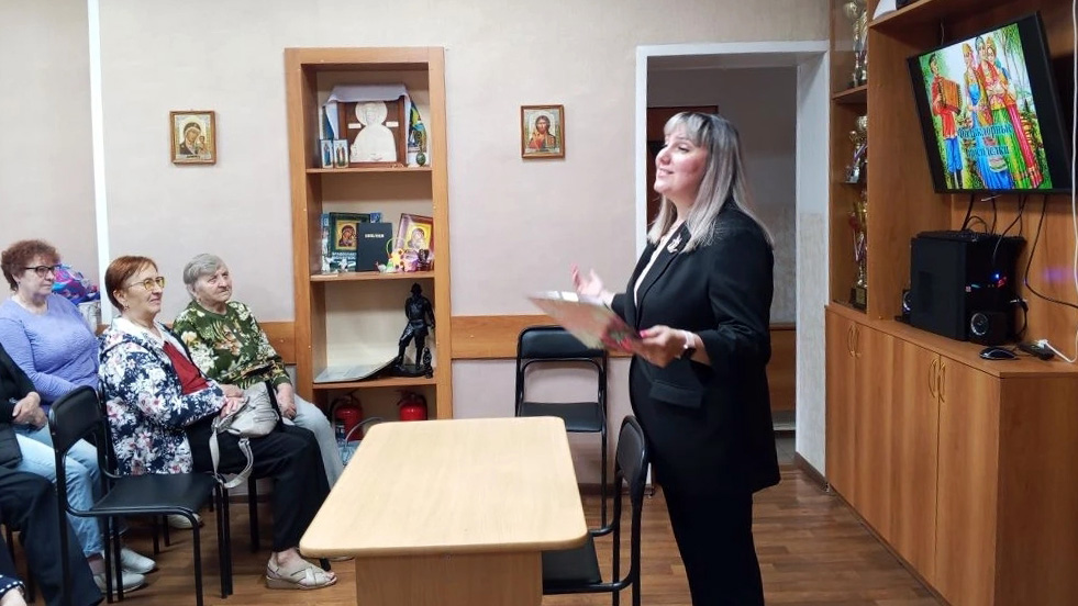 Женщина в чёрном костюме с папкой в руках стоит у стола и выступает перед рядом сидящими людьми в комнате с иконами на стене и включённым телевизором справа.