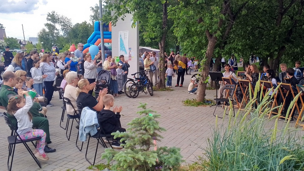 Уличное мероприятие в парке: справа сидит группа музыкантов с пультами, слева и по центру зрители, многие хлопают, вокруг деревья, детский надувной батут и много людей, гуляющих и наблюдающих.