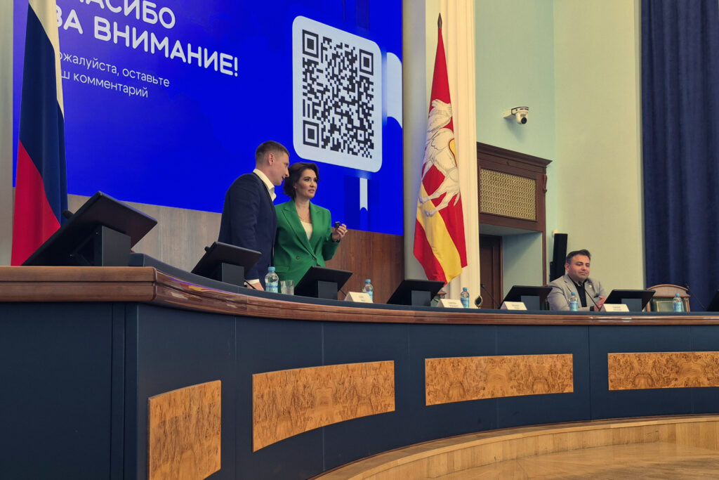 На президиуме перед большим синим экраном «Спасибо за внимание!» стоят мужчина в тёмном костюме и женщина в зелёном пиджаке, справа за столом сидит ещё один мужчина