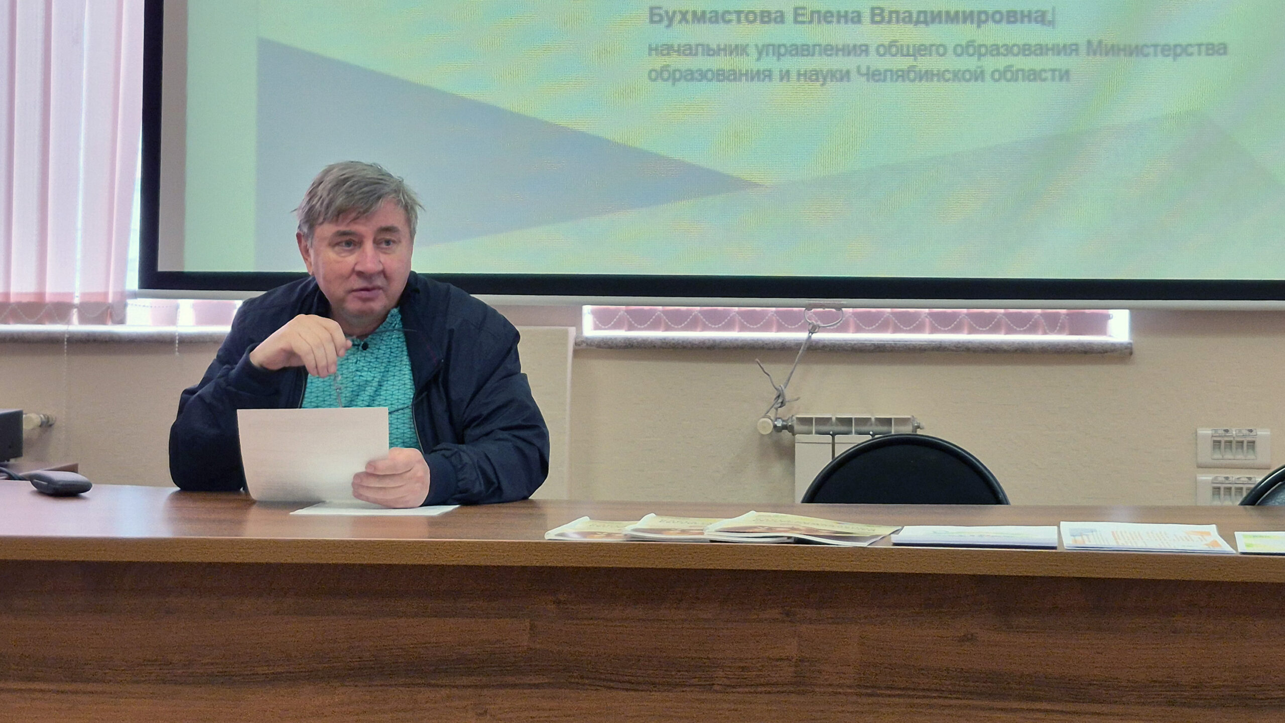 Мужчина сидит за столом с бумагами перед большим экраном и выступает, держа лист в руках.