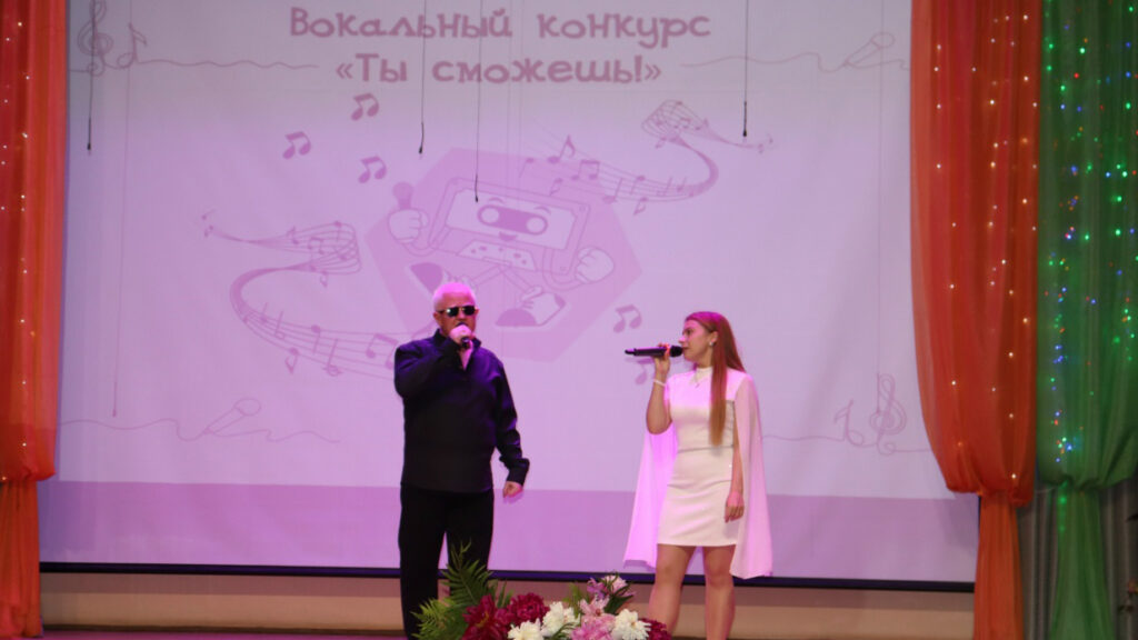 На сцене двое поют с микрофонами: мужчина в чёрной одежде и женщина в коротком белом платье с накидкой