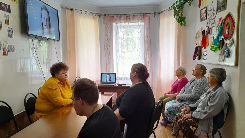 Комната, люди сидят за длинным столом и у стены, смотрят видеовыступление женщины на телевизоре и ноутбуке. На стенах фотографии и вязаные изделия, у окна светлые шторы.