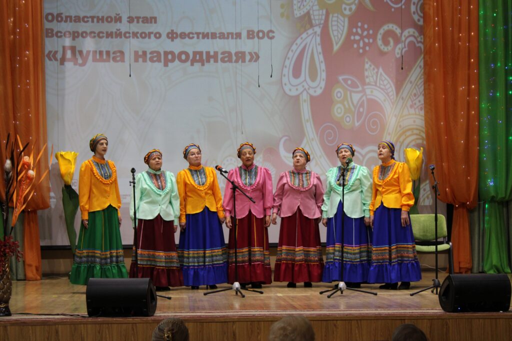 На сцене женский вокальный ансамбль из восьми человек в ярких народных костюмах: длинные юбки с орнаментом и разноцветные кофты, все в кокошниках.