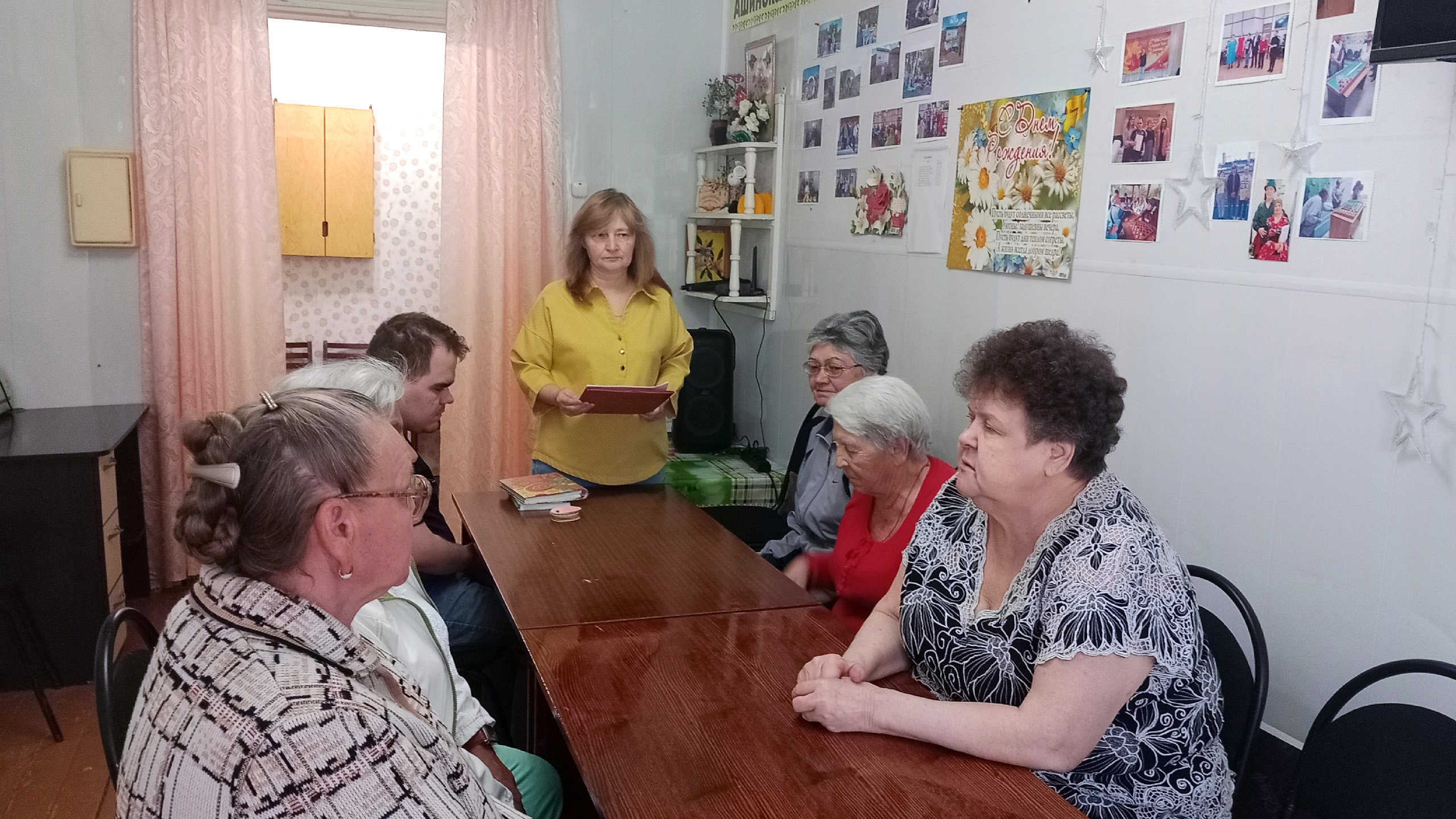 За длинным столом сидит группа пожилых людей и мужчина помоложе, а женщина в жёлтой блузке стоит у торца с папкой
