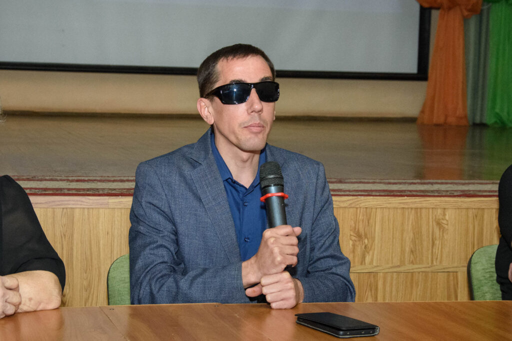 На фото мужчина в тёмных очках и синем пиджаке сидит за столом, держит микрофон; перед ним лежит смартфон, на заднем плане сцена.