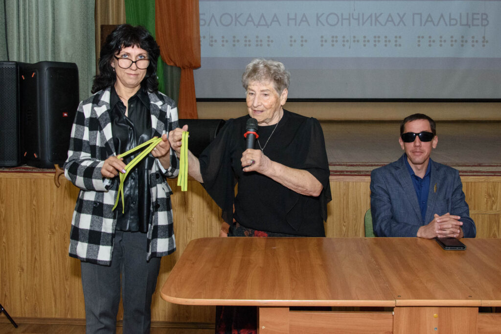На фото пожилая женщина с микрофоном что‑то вручает женщине в чёрно‑белом клетчатом пиджаке; справа за столом сидит мужчина в тёмных очках, перед ним смартфон.