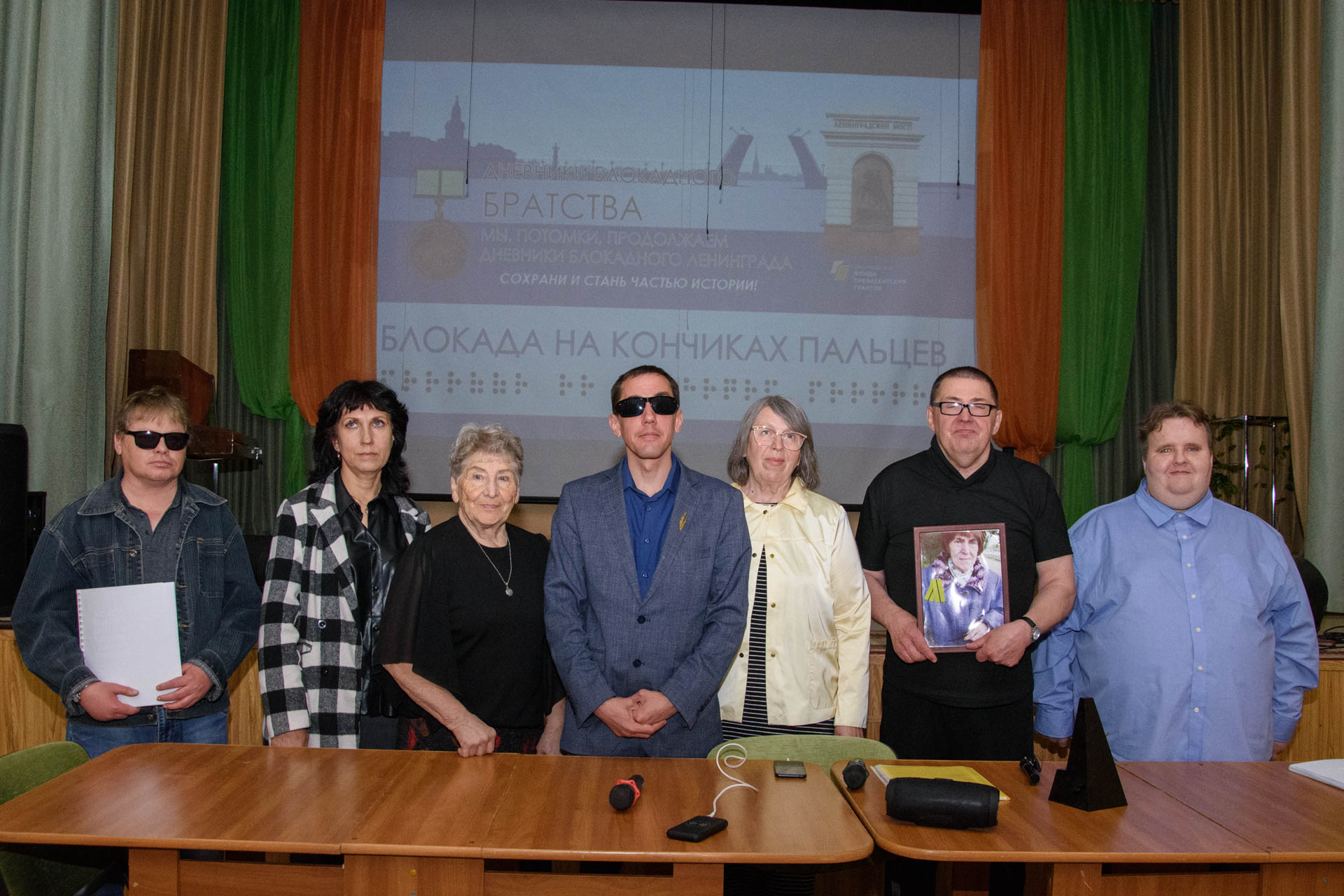 Семь человек стоят за столом в зале на фоне экрана с надписью «Блокада на кончиках пальцев»; один держит фотографию, перед ними на столе микрофоны и бумаги.