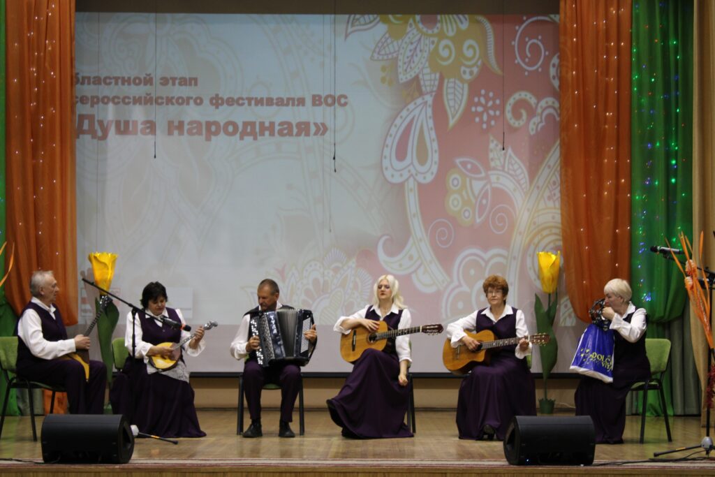 На сцене сидит ансамбль из шести человек в одинаковых фиолетово‑белых костюмах: играют на гитарах, аккордеоне и струнных инструментах
