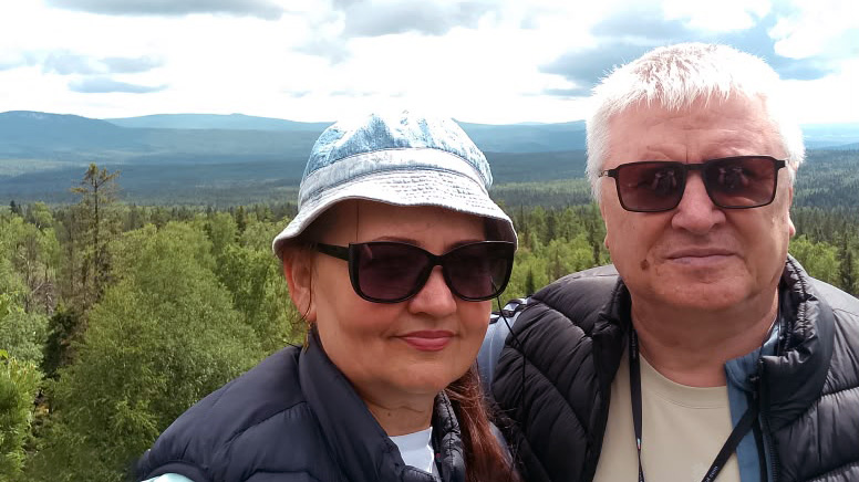 На переднем плане мужчина и женщина в тёмных куртках и солнцезащитных очках; у женщины светлая панама. На заднем плане зелёный лес и горы под облачным небом.