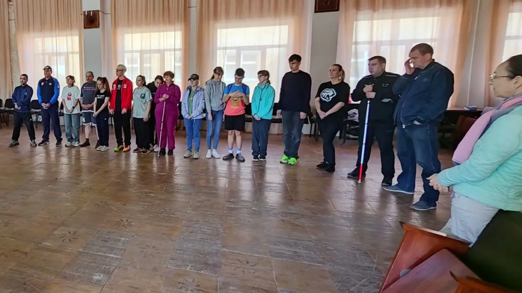 В большом зале стоит полукругом группа из примерно 15–16 человек, некоторые с тростями; похоже на общее построение или собрание.