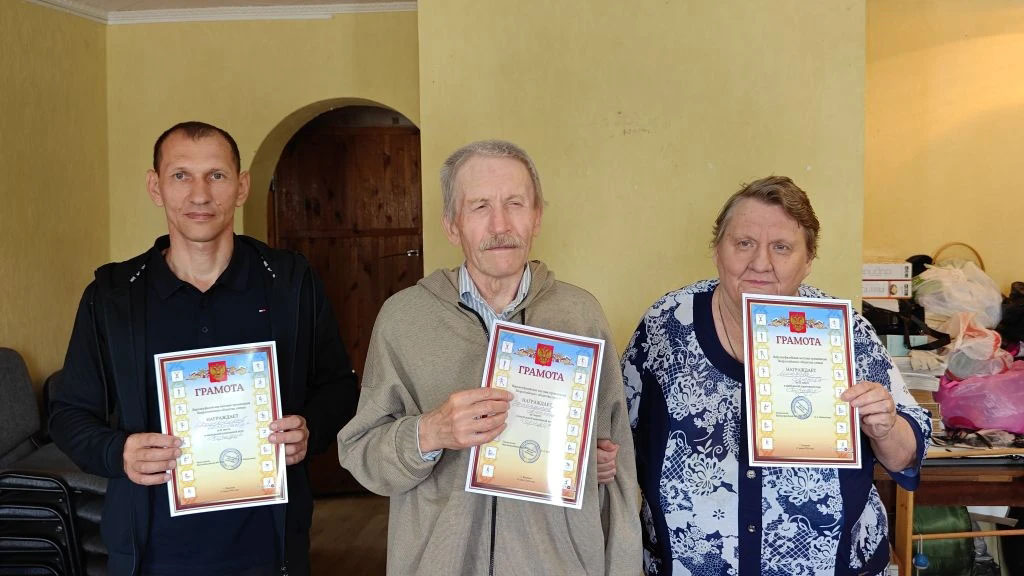 Трое людей (мужчина, пожилой мужчина и женщина) стоят в помещении и держат в руках грамоты с официальной символикой, позируя для фото.