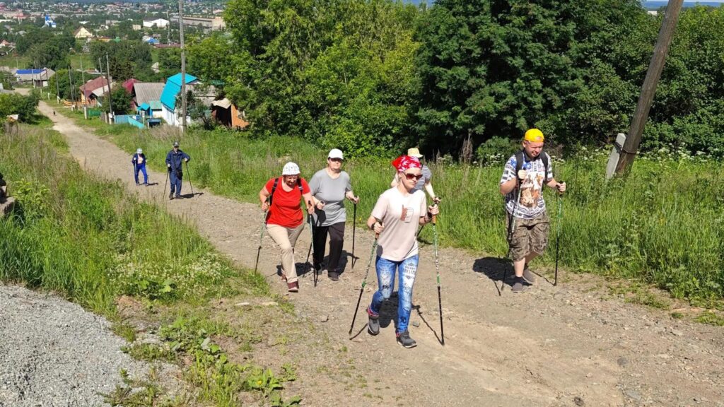 Группа людей с треккинговыми палками поднимается по грунтовой дороге в гору. Вокруг зелёная трава и деревья, на заднем плане видны дома посёлка и другие участники, идущие позади.
