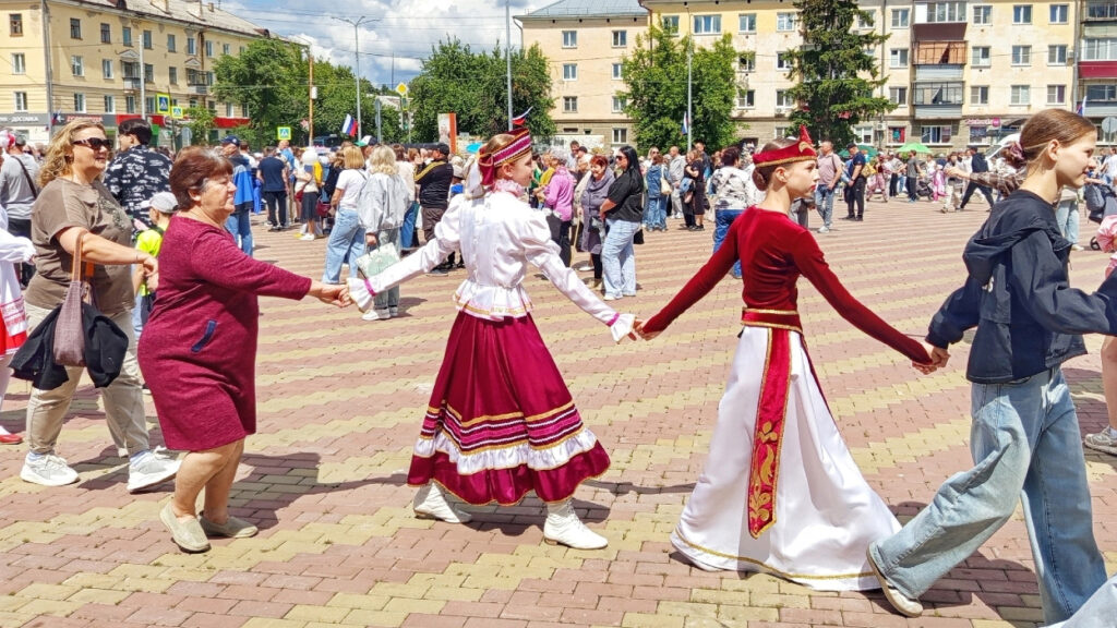 Люди на городской площади водят хоровод, держась за руки. Среди них девушки в ярких национальных костюмах бордово‑белых цветов и обычные горожане в повседневной одежде