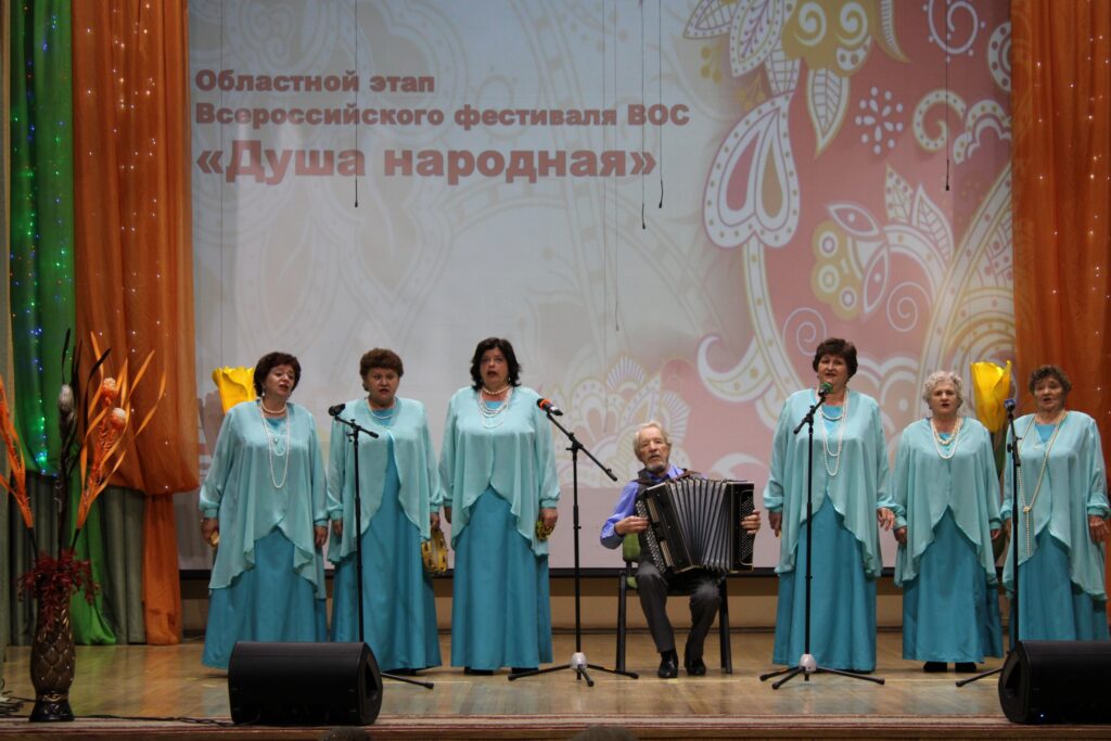 На сцене сидит ансамбль из шести человек в одинаковых фиолетово‑белых костюмах: играют на гитарах, аккордеоне и струнных инструментах