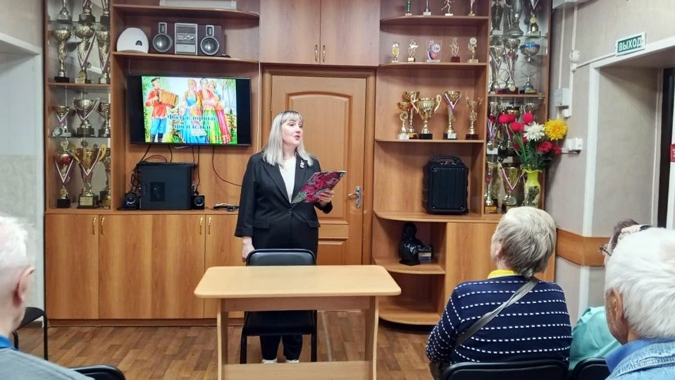 Женщина в чёрном костюме стоит перед столом и читает по папке перед группой людей в комнате с витринами, полными кубков; на стене за ней включён телевизор.