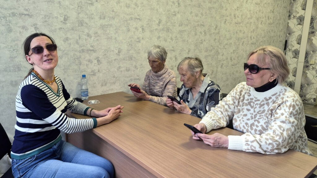 За столом сидят четыре женщины.
Три пожилые женщины справа изучают смартфоны.
Слева женщина помоложе в полосатой кофте и тёмных очках, повернута лицом к камере.