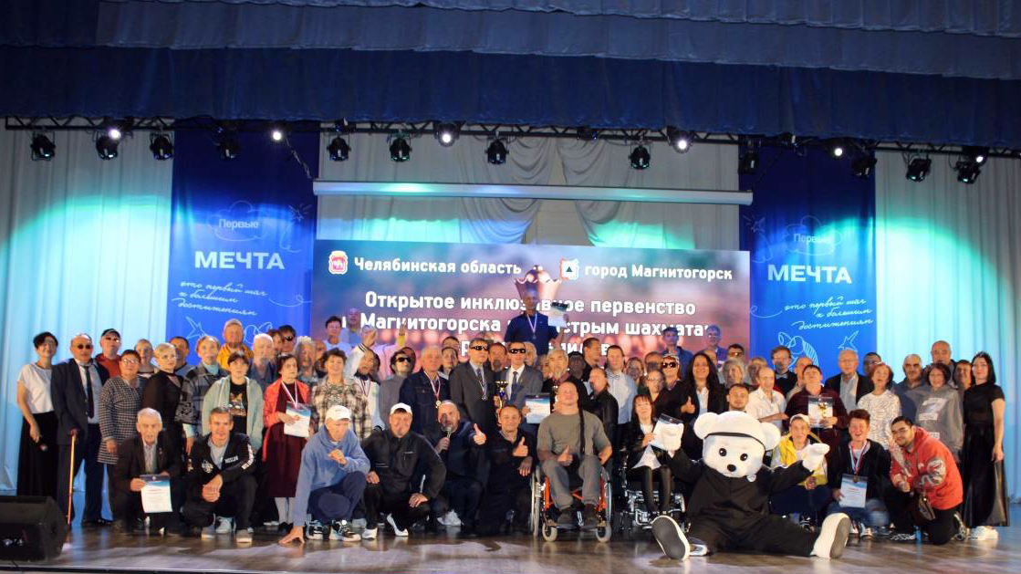 Групповое фото на сцене: много участников турнира по быстрым шахматам в Магнитогорске на фоне баннера с надписью о открытом инклюзивном первенстве, спереди сидит человек в костюме белого медведя.