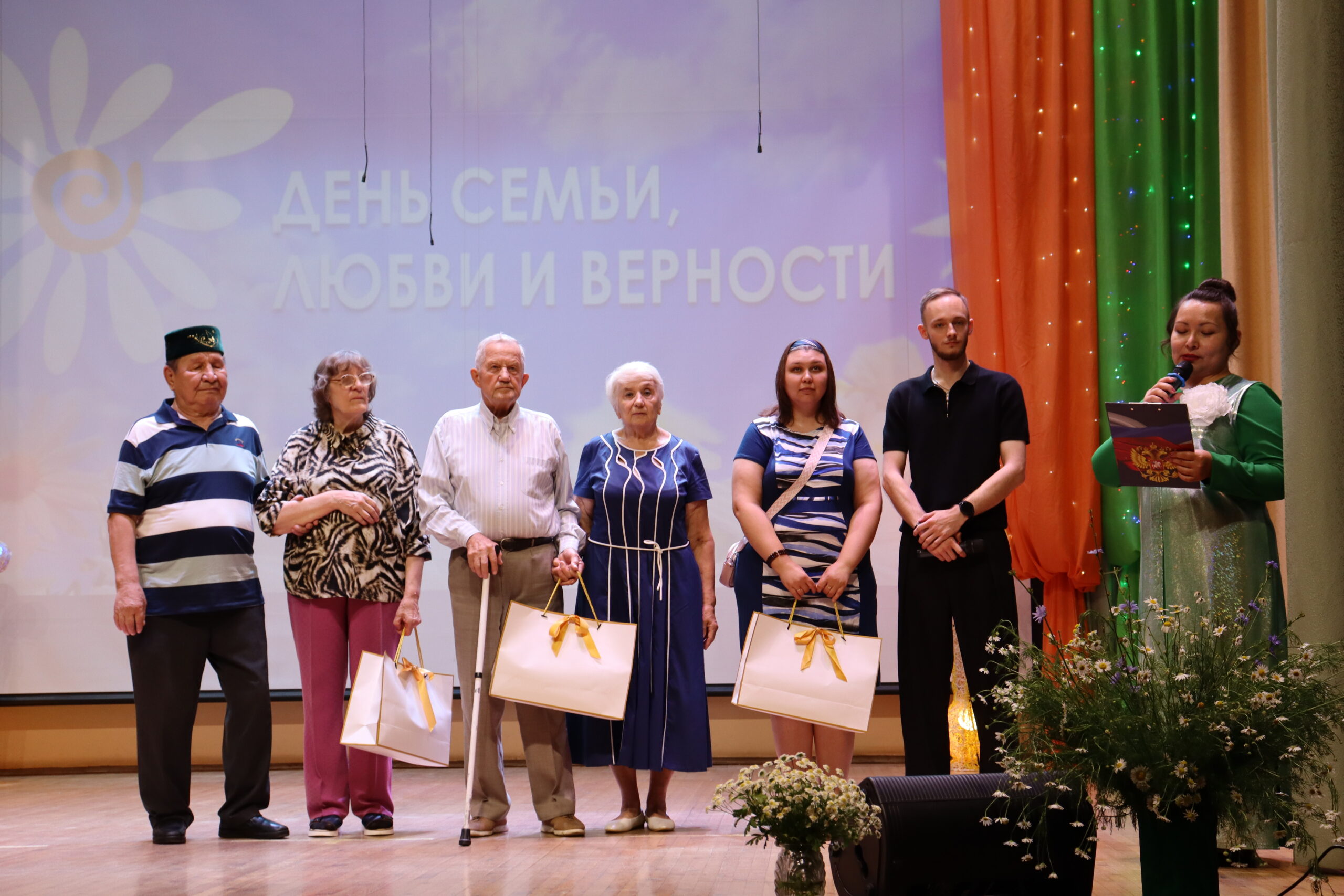 На сцене шесть человек с подарочными пакетами на фоне надписи «ДЕНЬ СЕМЬИ, ЛЮБВИ И ВЕРНОСТИ». Справа ведущая в зелёном платье говорит в микрофон. Спереди на сцене стоят букеты полевых цветов с ромашками.