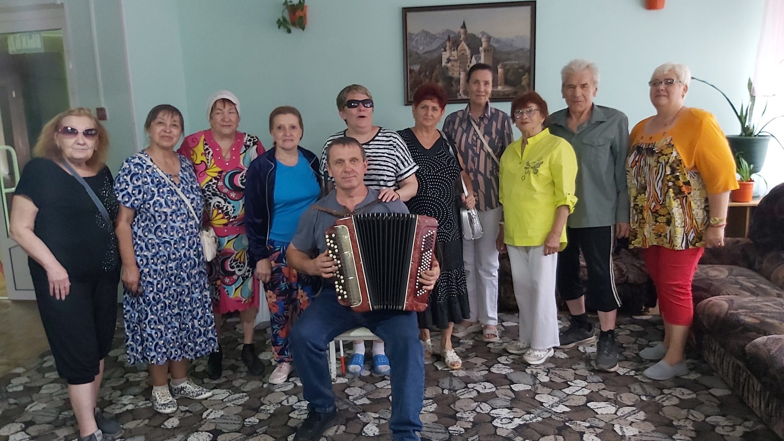 На фото группа из 12 пожилых людей в комнате, в центре мужчина сидит с аккордеоном, остальные стоят полукругом позади него, улыбаются; за ними на стене картина с замком.