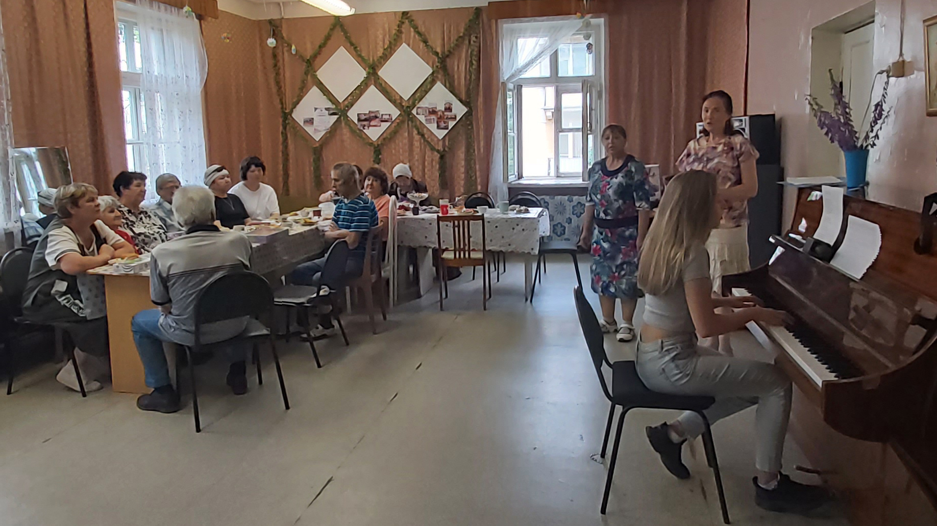 В комнате за двумя накрытыми столами сидит группа людей, а у пианино справа молодая женщина играет, рядом стоят две женщины, похоже, поют; это напоминает дружеское застолье с музыкальным выступлением.