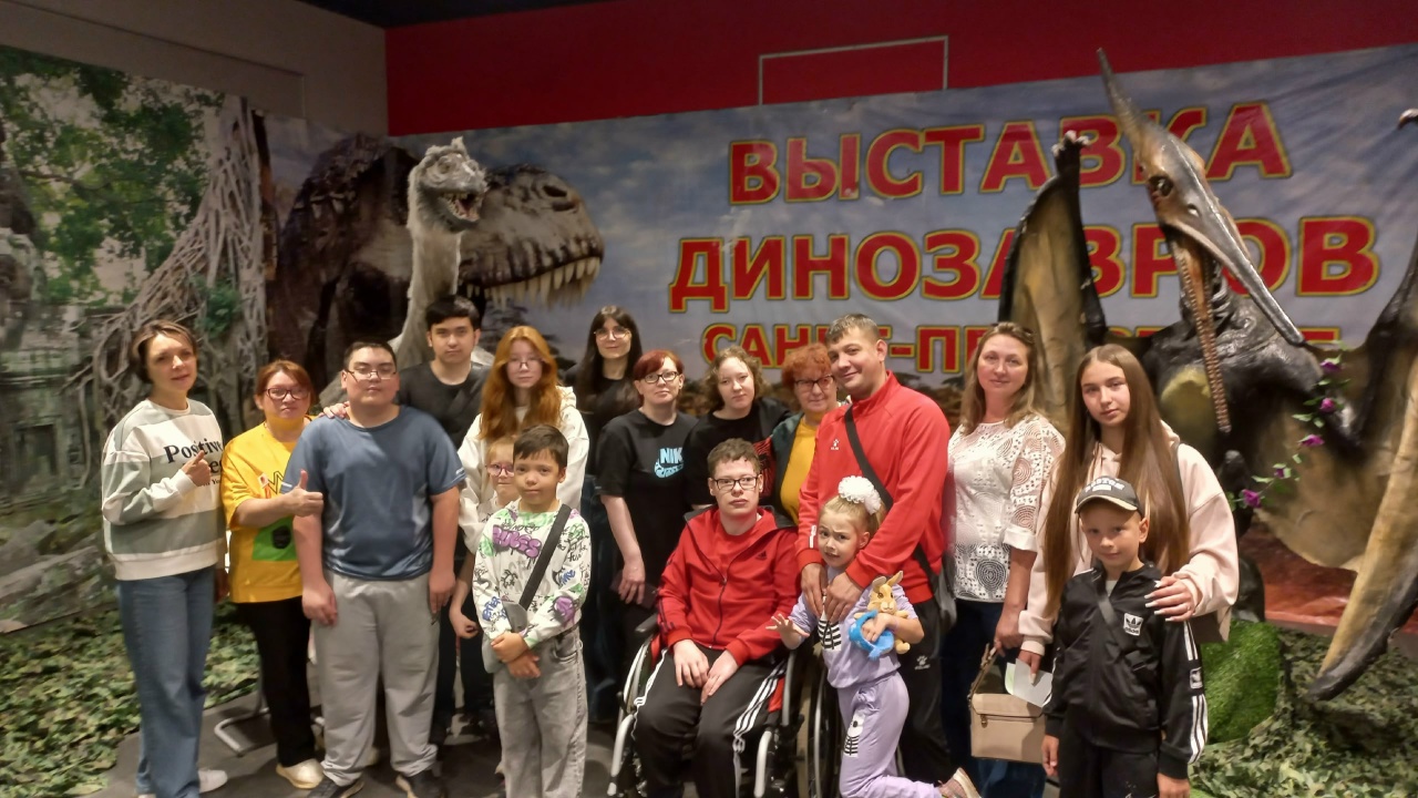 Группа детей и взрослых позирует для фото на фоне баннера «Выставка динозавров Санкт‑Петербург» и больших фигур динозавров.