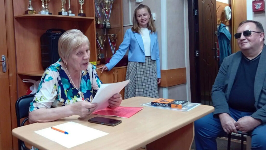 Женщина за столом читает лист бумаги, перед ней лежат телефон и ручка.
Напротив сидит мужчина в тёмных очках, сзади у шкафа с кубками стоит женщина в голубом жакете.