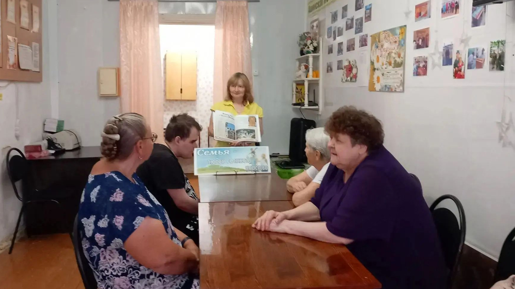 Несколько человек сидят за столом, а женщина в жёлтой блузке у торца стола показывает открытую книгу; перед ней стоит табличка с надписью «Семья – это маленькое царство».