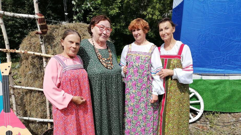 Четыре женщины в русских сарафанах стоят на улице у стога сена и декораций, смотрят в камеру и улыбаются.