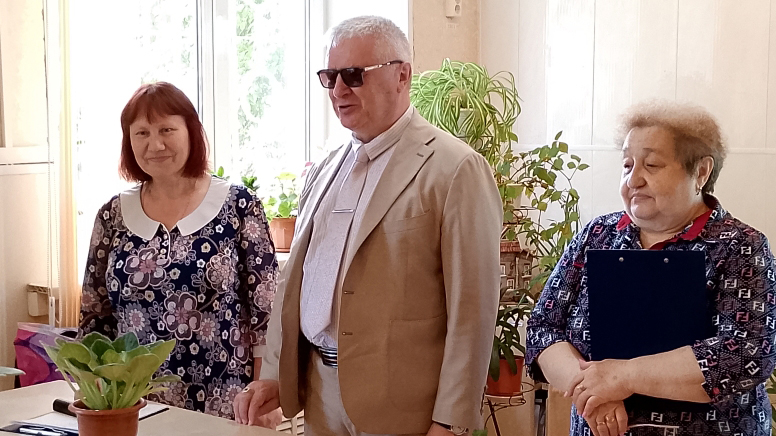 На фото трое взрослых людей в комнате у окна с комнатными растениями.