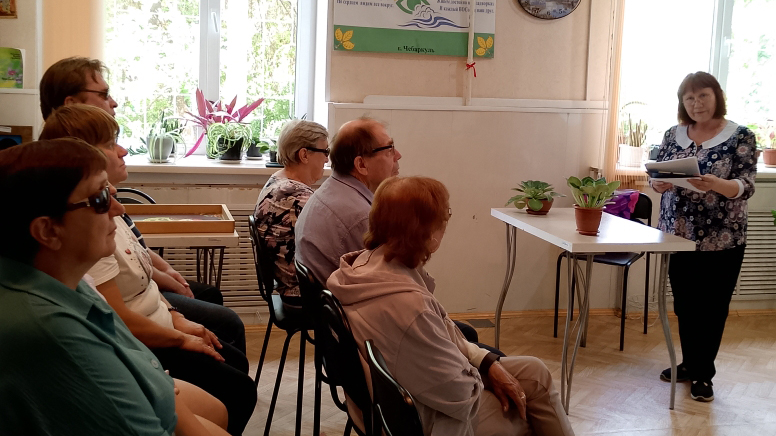 На фото несколько взрослых сидят в ряд на стульях, повернувшись к женщине, которая стоит справа у стола и что‑то читает с бумаг.