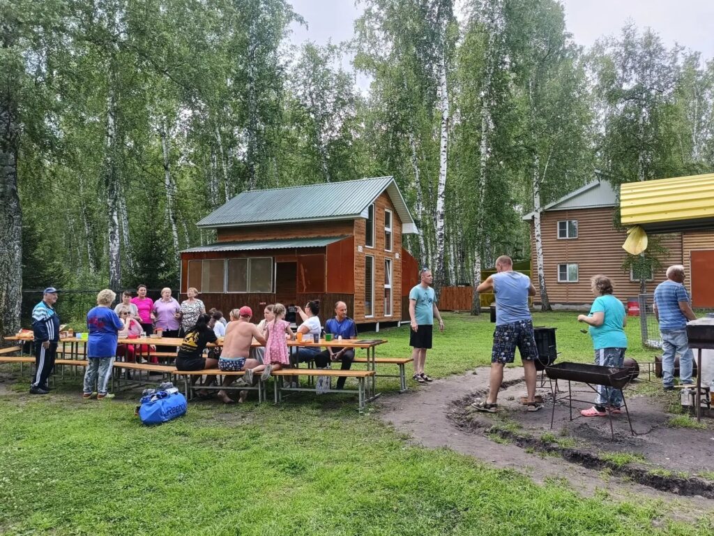 Группа людей на базе отдыха: часть сидит за длинными деревянными столами, другие стоят у мангалов и общаются; на заднем плане видны деревянные домики среди берёз.