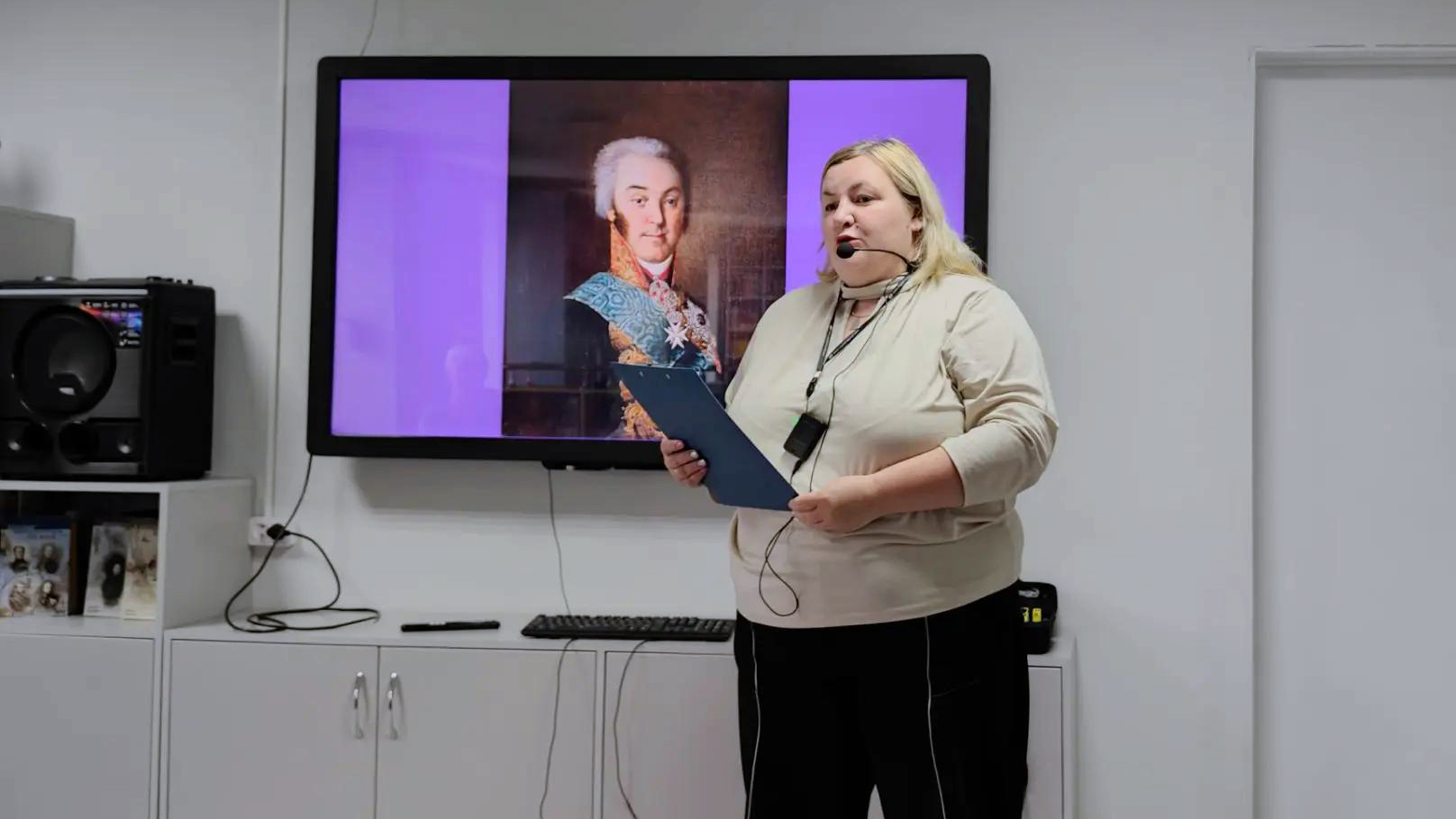 Женщина в светлой кофте и темных брюках стоит в комнате с микрофоном и папкой, читает доклад. За ней на большом экране висит слайд с портретом мужчины в парадной одежде. Справа белая дверь, слева музыкальный центр на тумбе.